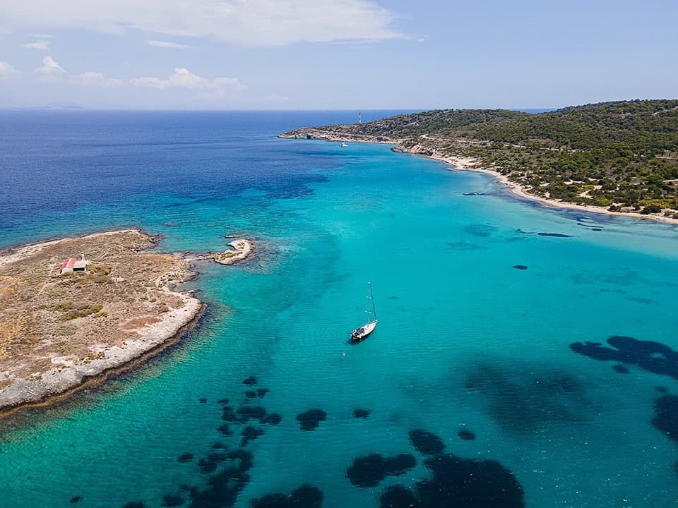 Aerial view of a sailing yacht anchored alone in a turquoise Greek bay beside a low islet