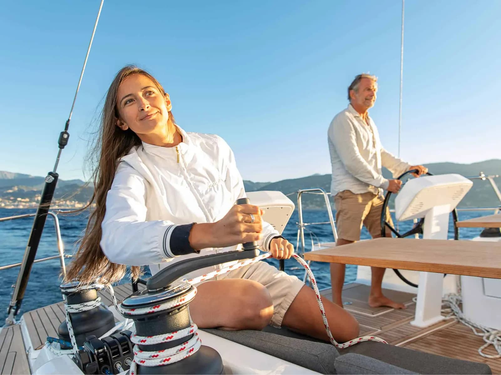 A woman working a winch while a man helms a sailing yacht