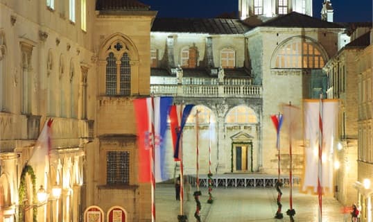 Luža square in Dubrovnik at night, lined with flags and lit stone facades