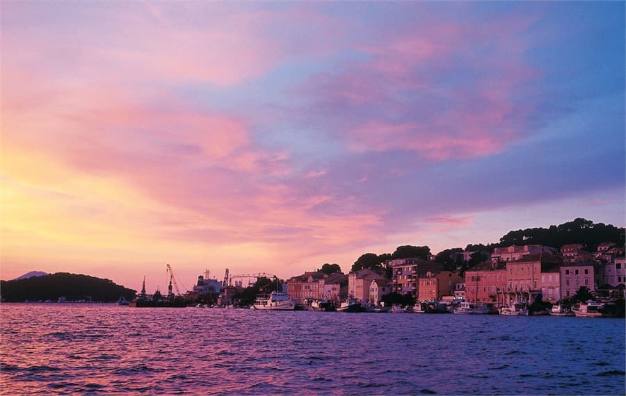 The waterfront of Mali Lošinj at sunset, pastel houses and moored fishing boats under a pink and violet sky
