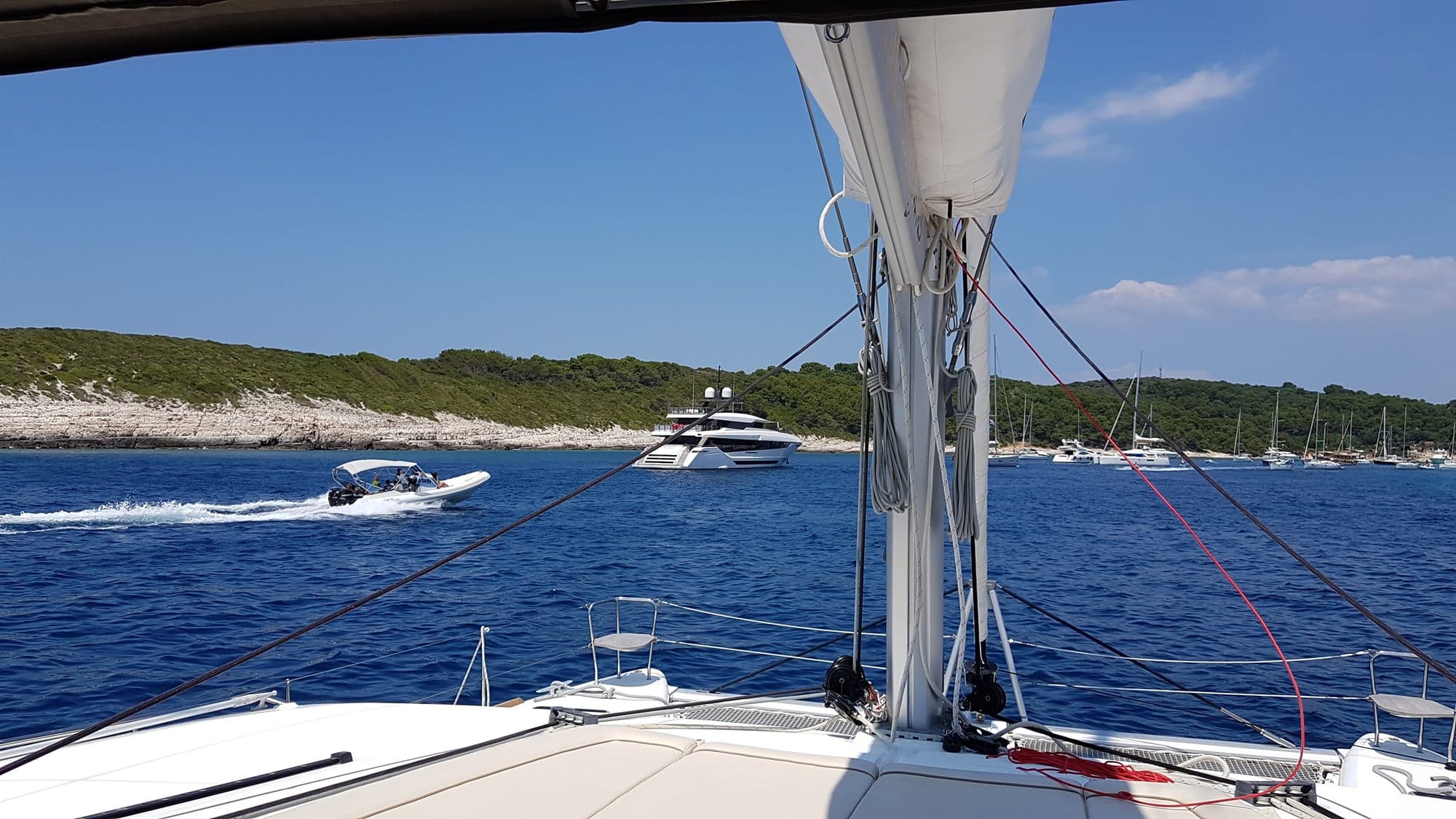 A white motor yacht lying off a pine-covered Croatian island with a tender running past, seen from the foredeck of another boat at anchor
