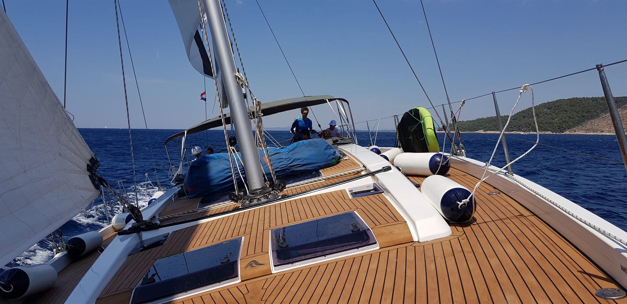 Teak foredeck of a cruising yacht heeling gently, fenders and a spinnaker bag stowed against the guardrail, deep blue water and a wooded island beyond