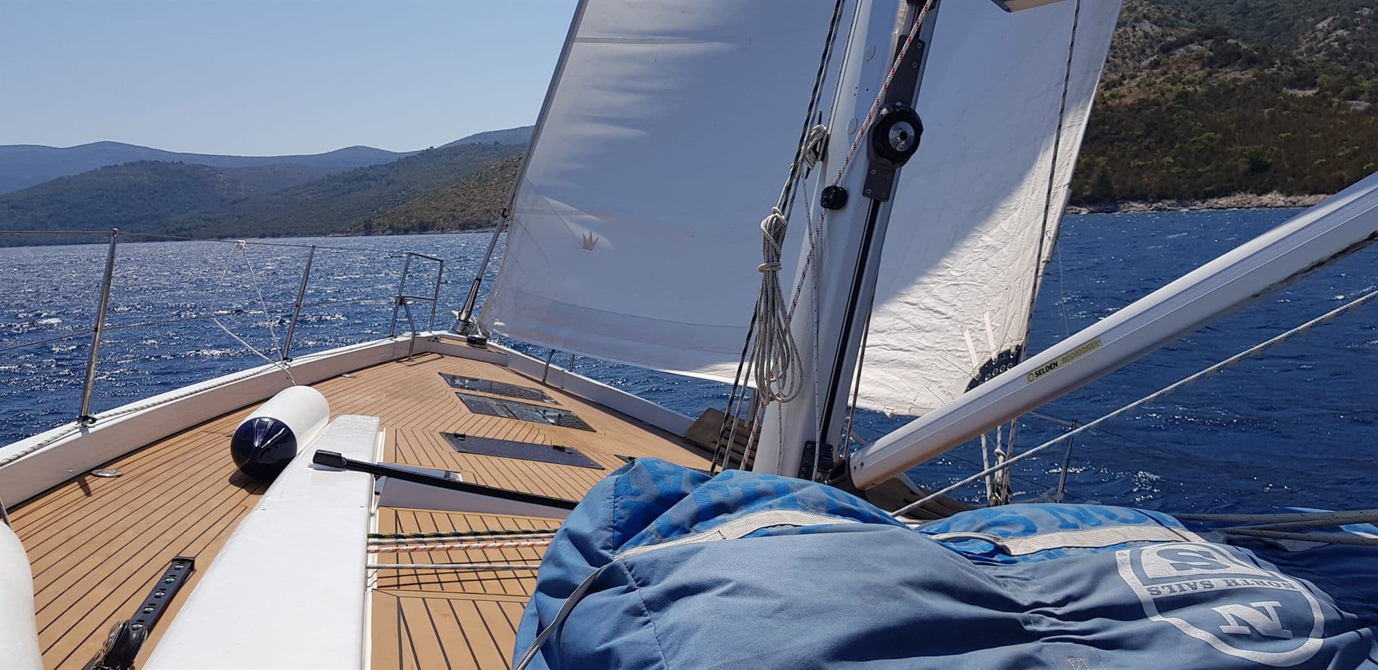 The foredeck of a sailing yacht under way in Croatia, genoa drawing and teak side deck running forward, with a bare Dalmatian headland a mile off the bow