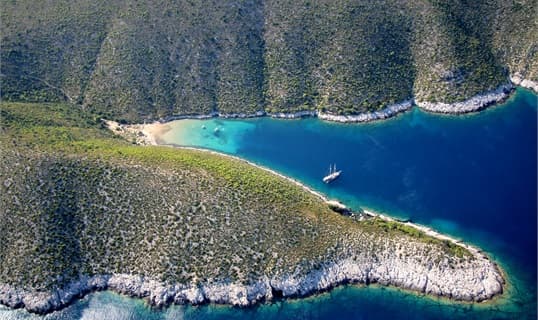 Aerial view of a yacht anchored alone in a deep blue bay on Vis, below steep scrub-covered slopes