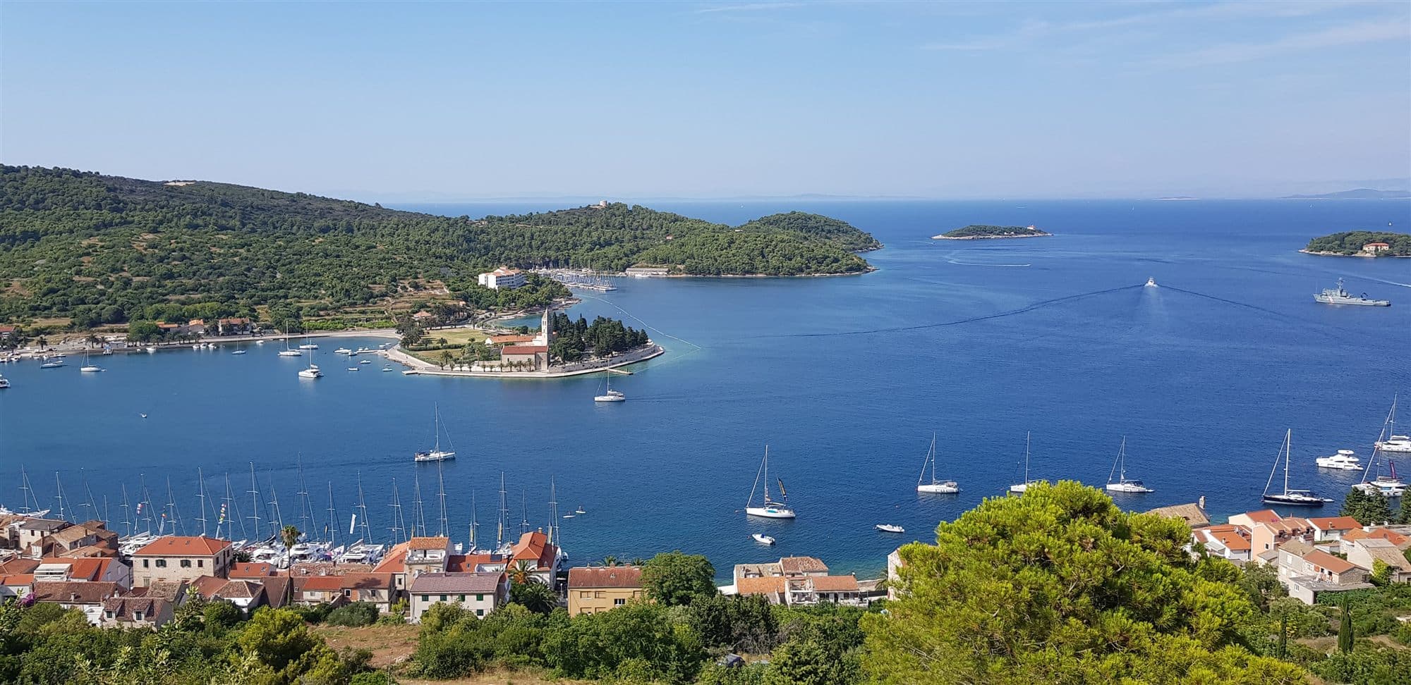Vis Town seen from the hillside above it: the harbour full of masts, yachts at anchor across the bay, and the Franciscan monastery on its wooded point