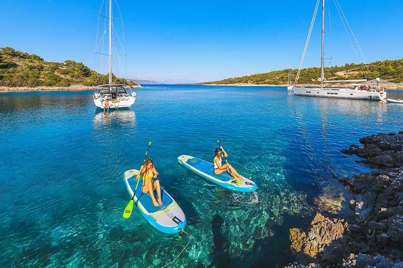 Two paddleboarders in a clear Croatian bay with sailing yachts at anchor